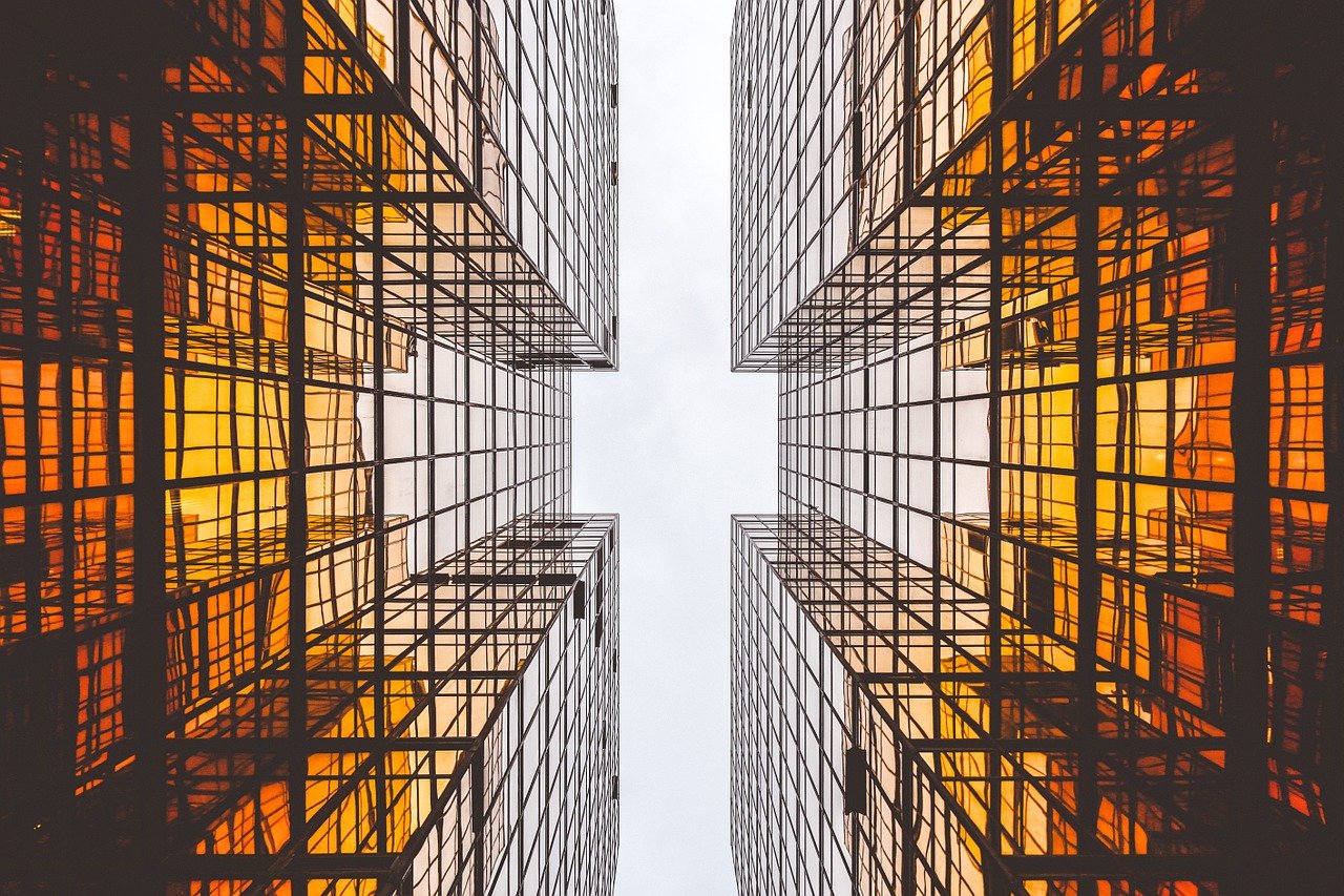 A symmetrical photograph of what appears to be two identical buildings. The perspective of the photograph is from the middle of the two buildings, looking up towards the sky. There is a reflection of the opposite building in each building's windows.